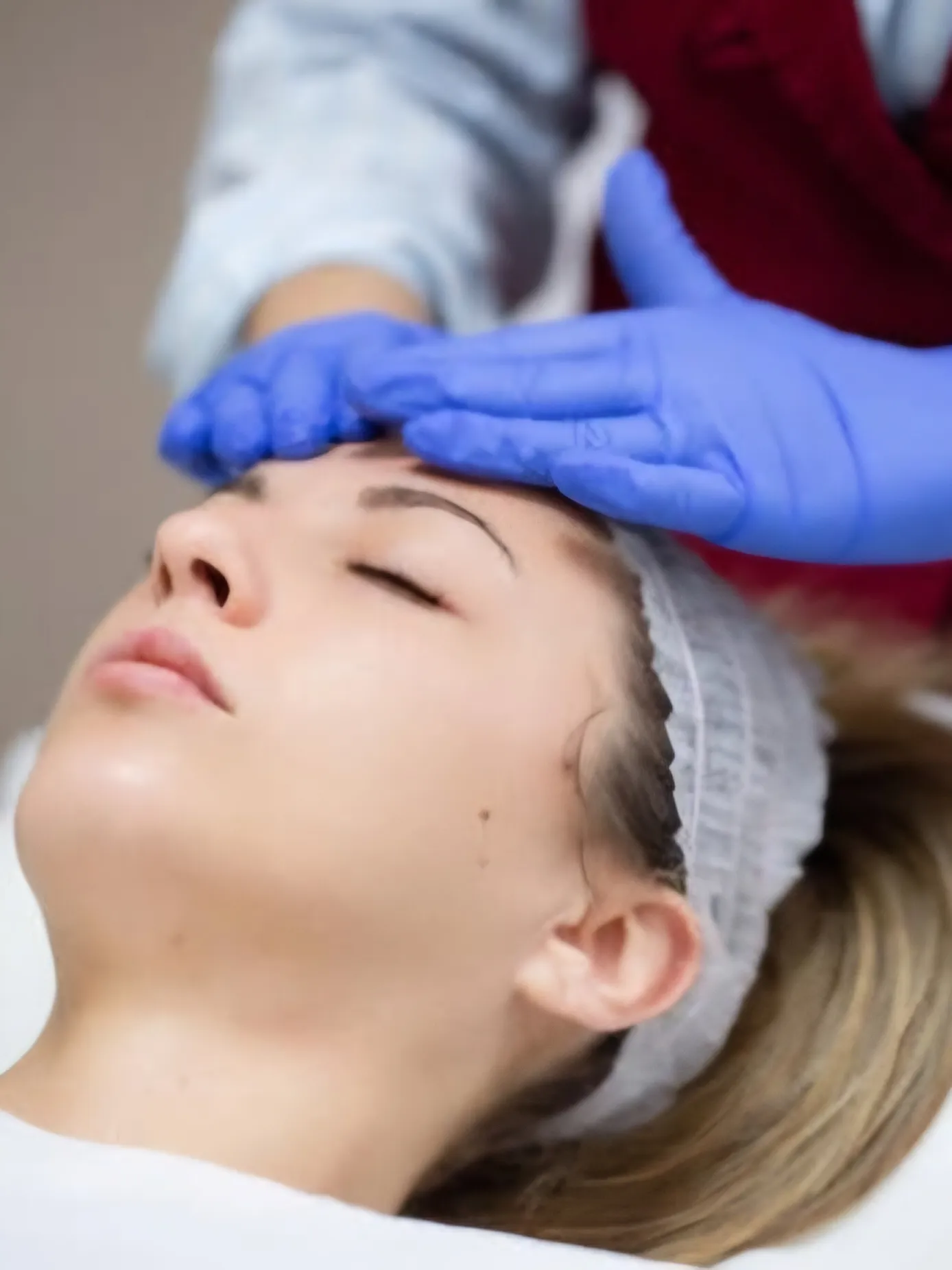 Paziente distesa a occhi chiusi durante un trattamento viso, con le mani del medico in guanti sulla fronte
